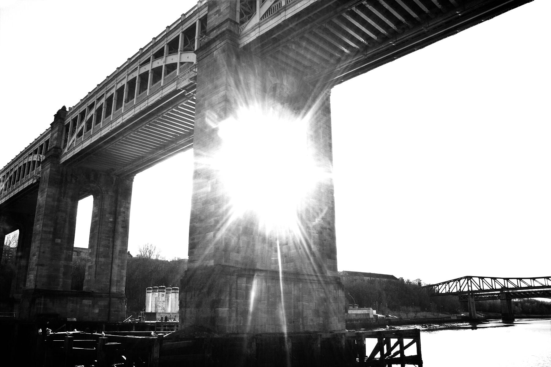 In black and white a tall sandstone bridge, with industrial construction on top for transport, Light shining through one of the supporting legs over a river.