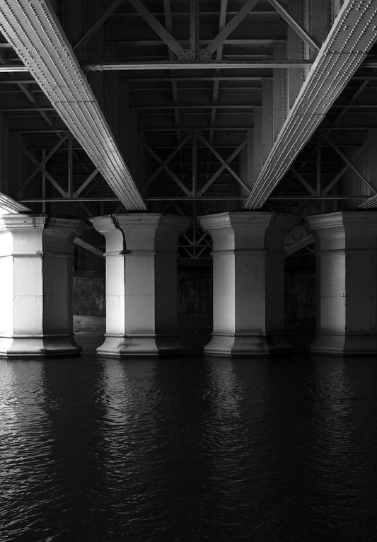 In the centre four industrial iron support pillars presented in black and white holding an iron bridge up on top of a body of water in the bottom half