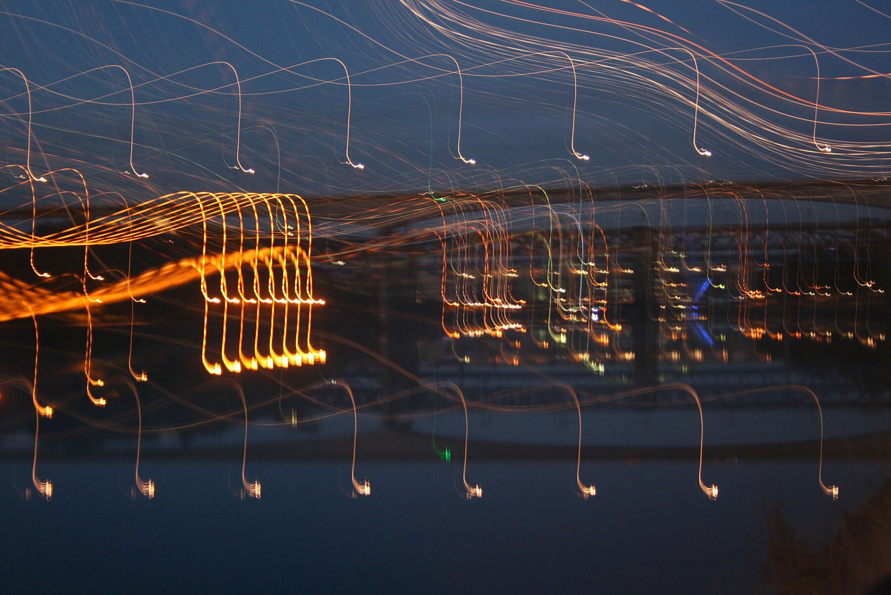 A cityscape over the water after sunset, bridges in the background, and lights reflecting off the water and made into light trails throughout the image.