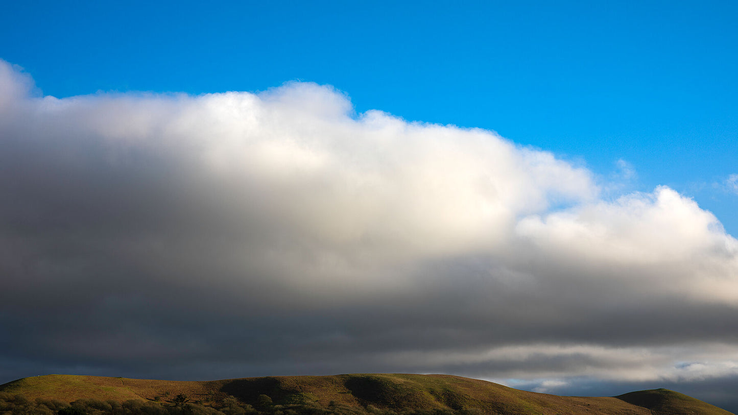 Scenic view of a rolling, washed-out green and orange hill with a large grey cloud hovering above it, set against a vibrant blue sky.