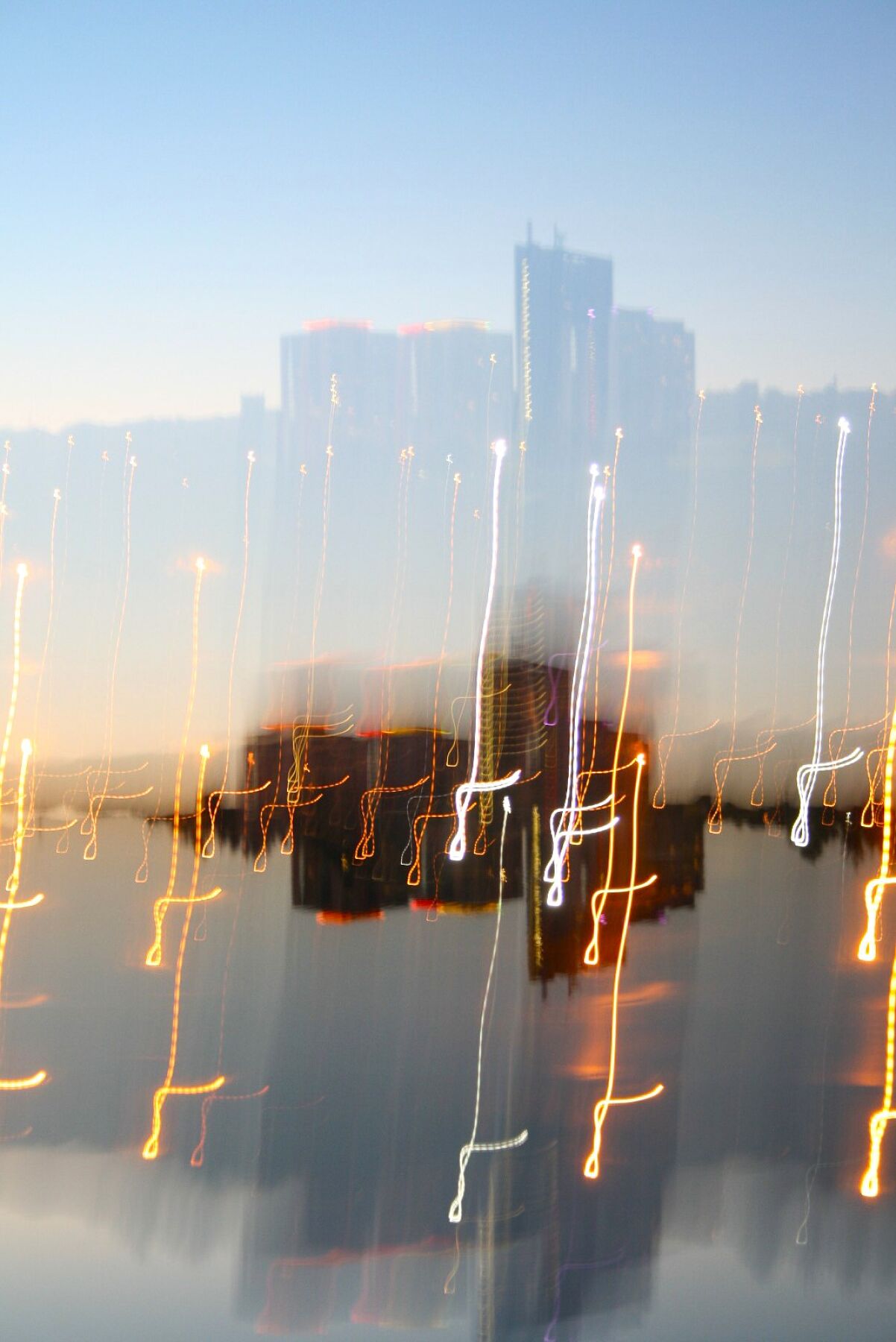 Abstract light play, buildings in the distance photographed and distorted to create light trails over a body of water just before dusk.