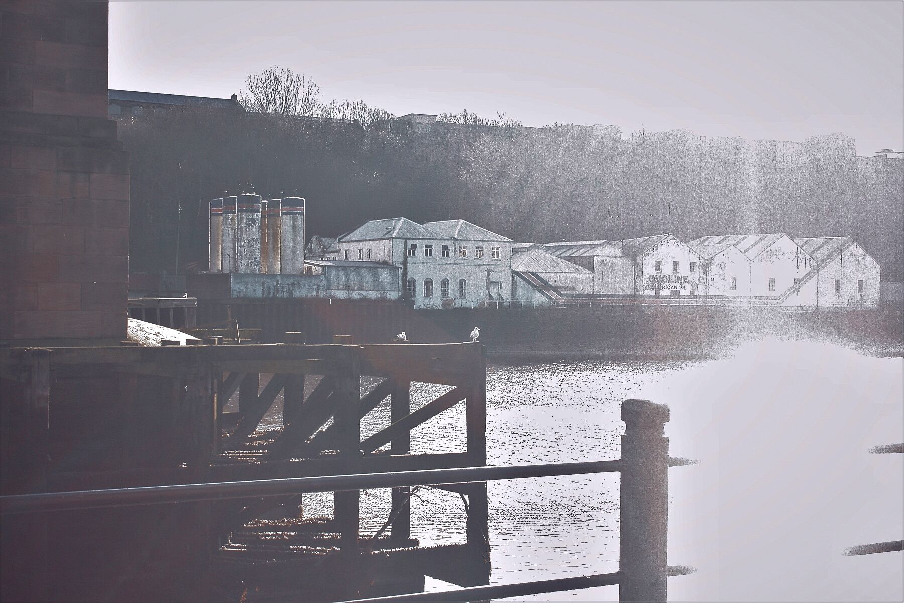 Architecture is now demolished, showing an abandoned lubricant factory across a river with the edge of a sandstone bridge support and wooden post in the foreground