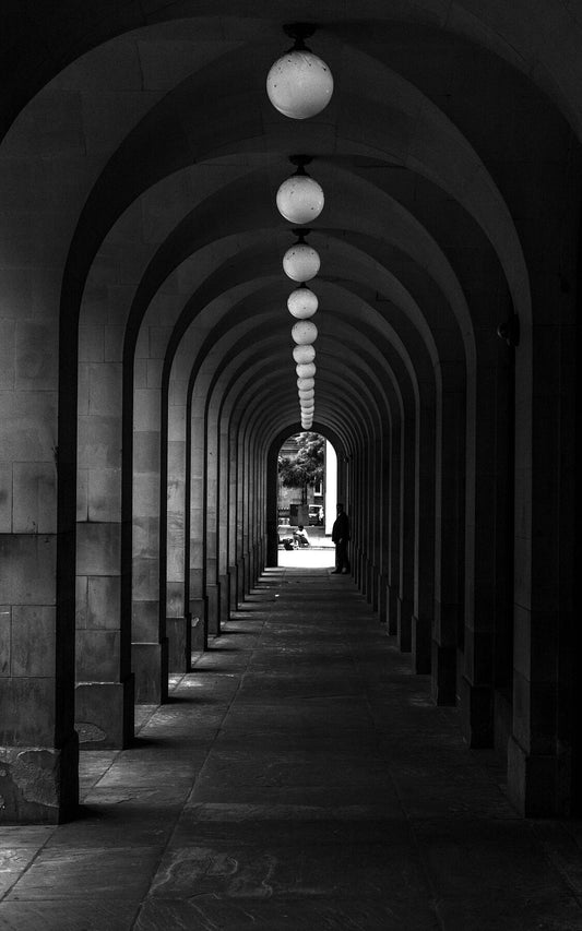 Passage. Arches adorned with hanging orb lights, a figure standing at the end, leading to a sunlit street with a seated individual and a tree in the background.